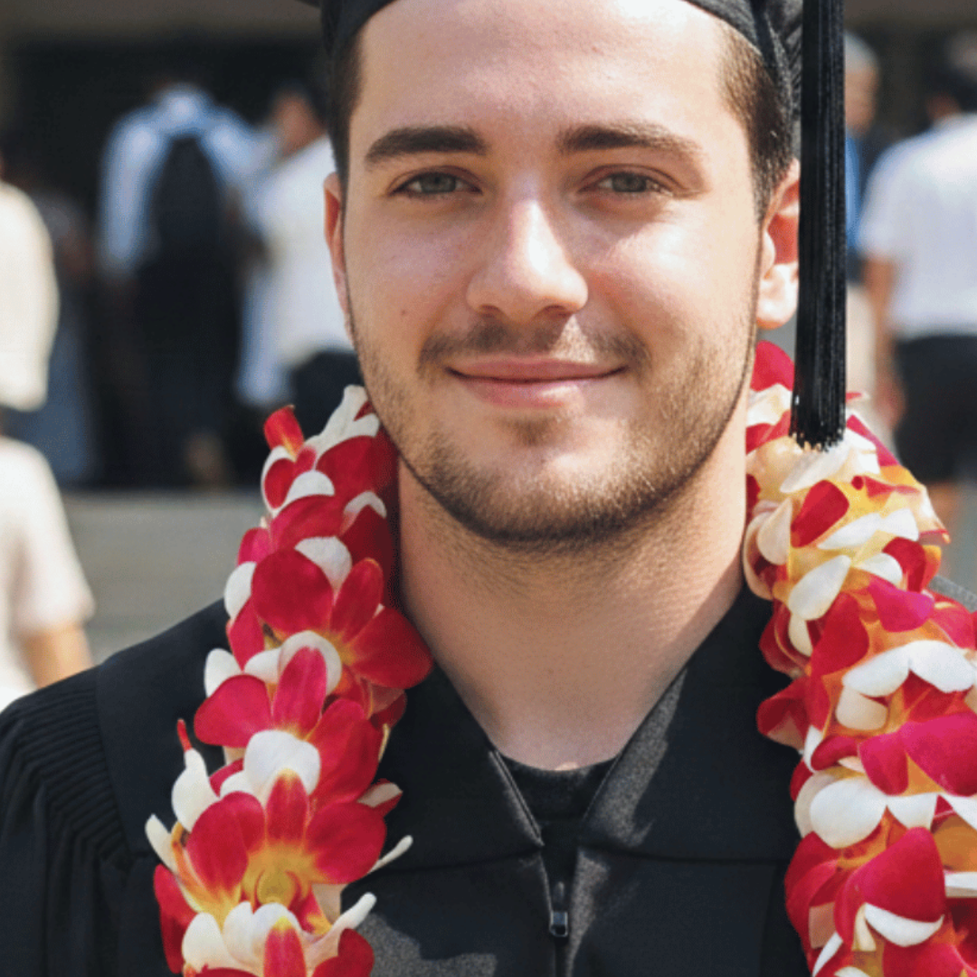 Graduate wearing a black cap and gown with a red and white lei, standing outdoors.